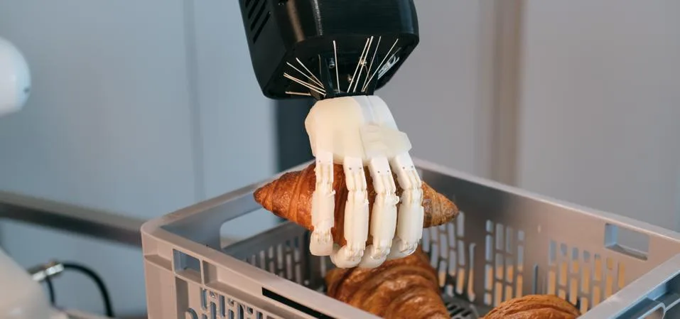 A white tendon-driven humanoid robotic hand lifting a croissant out of a crate.