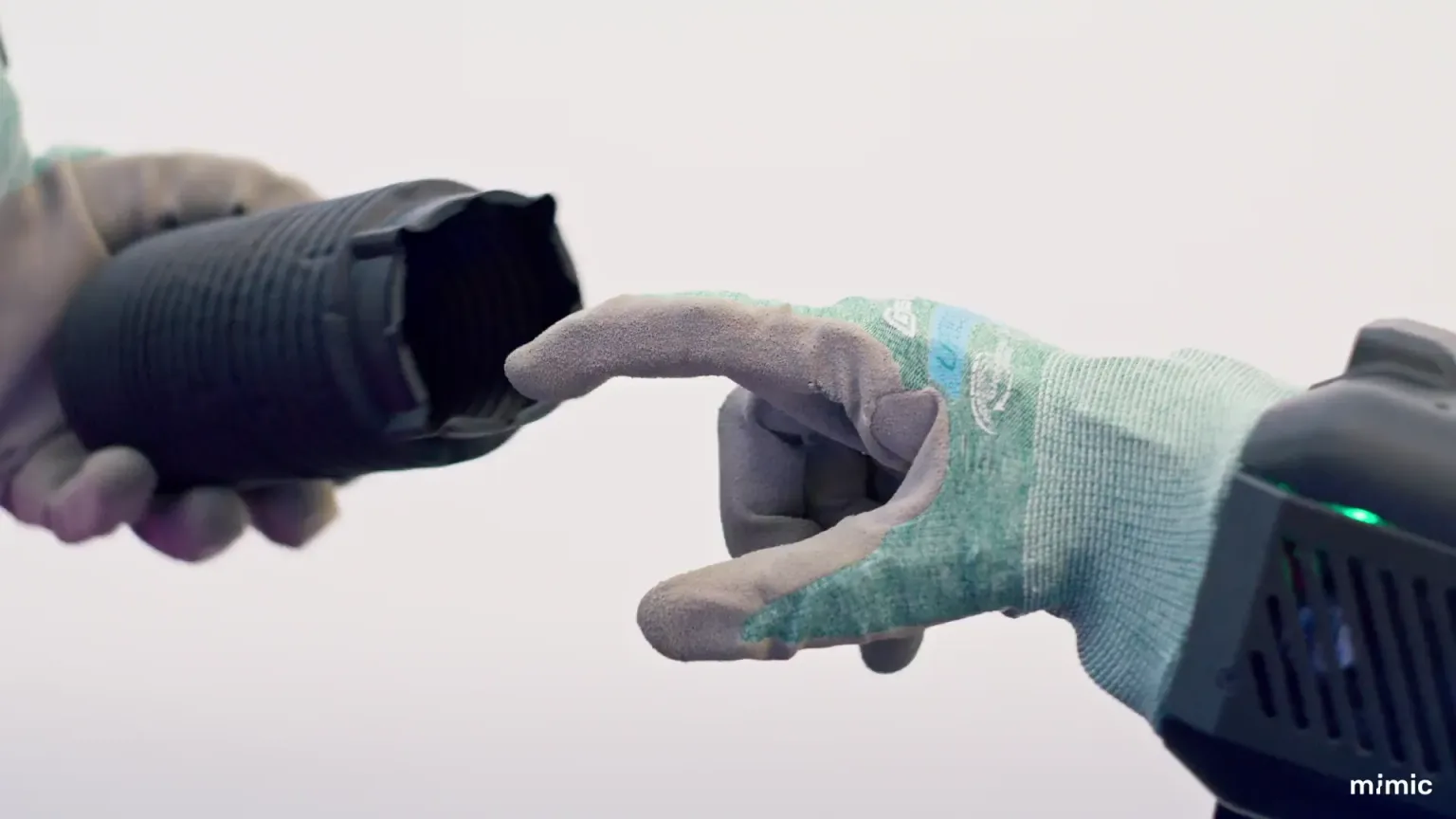 A mimic robotic hand in a glove reaches toward a person wearing a work glove in an industrial setting.