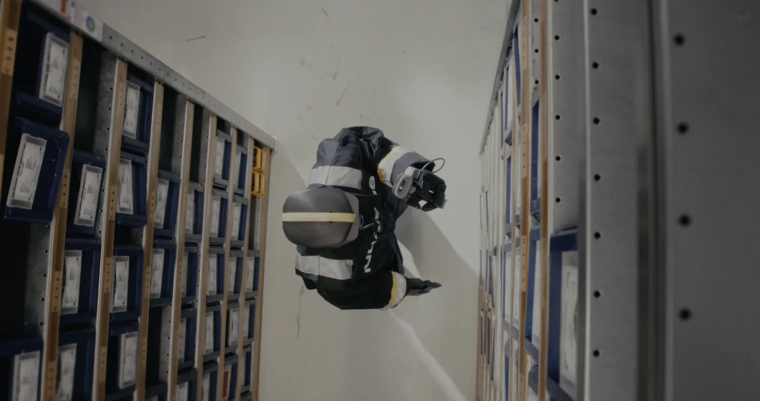 An overhead view of a Nucleus humanoid robot reaching toward parts bins between storage racks.
