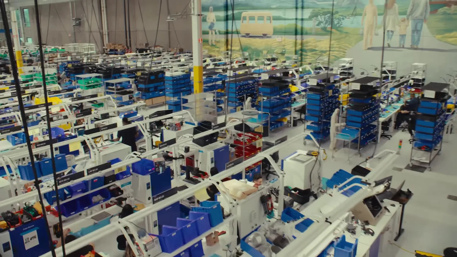 A wide view of 1X's NEO factory floor with rows of workstations, parts bins and humanoid robots.