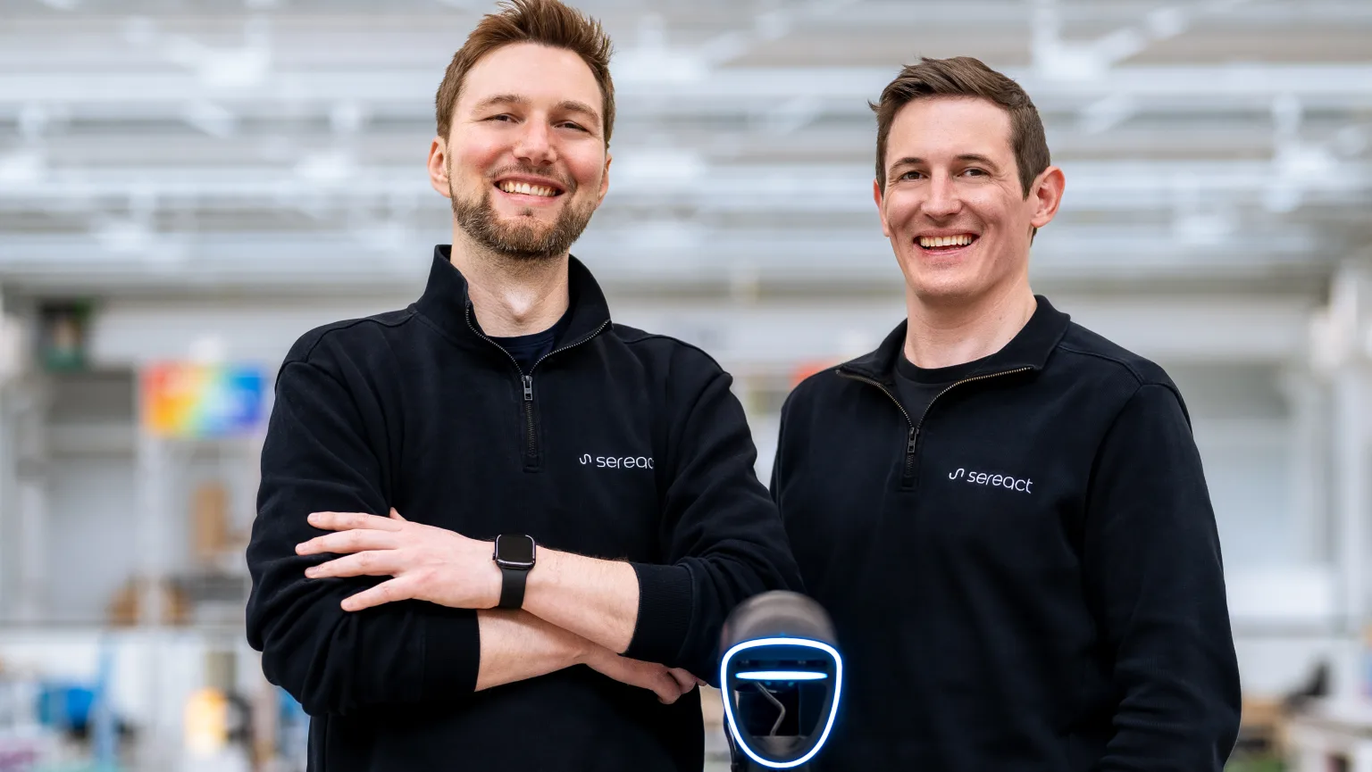 Sereact co-founders Ralf Gulde and Marc Tuscher standing behind a Sereact humanoid robot in a workshop.