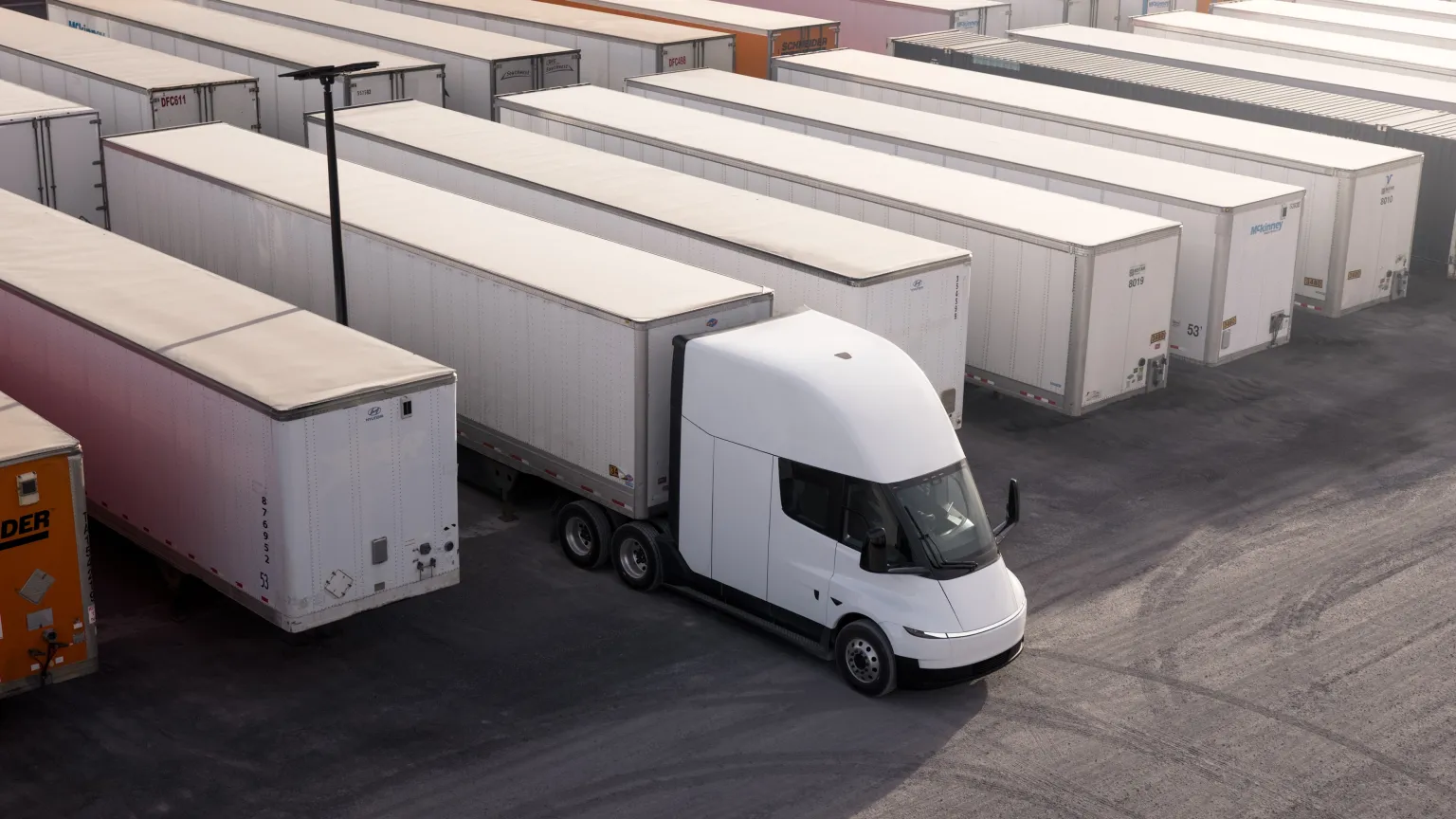 A white Tesla Semi maneuvering among rows of freight trailers at a logistics yard.